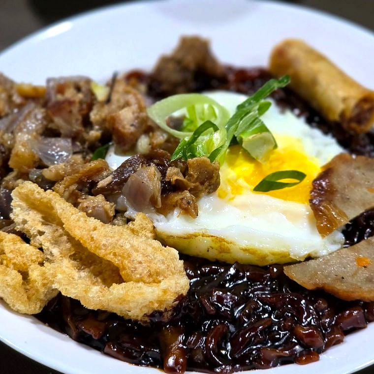 Black Guisado Tamis-Anghang — dark savory noodles topped with egg and chicharon